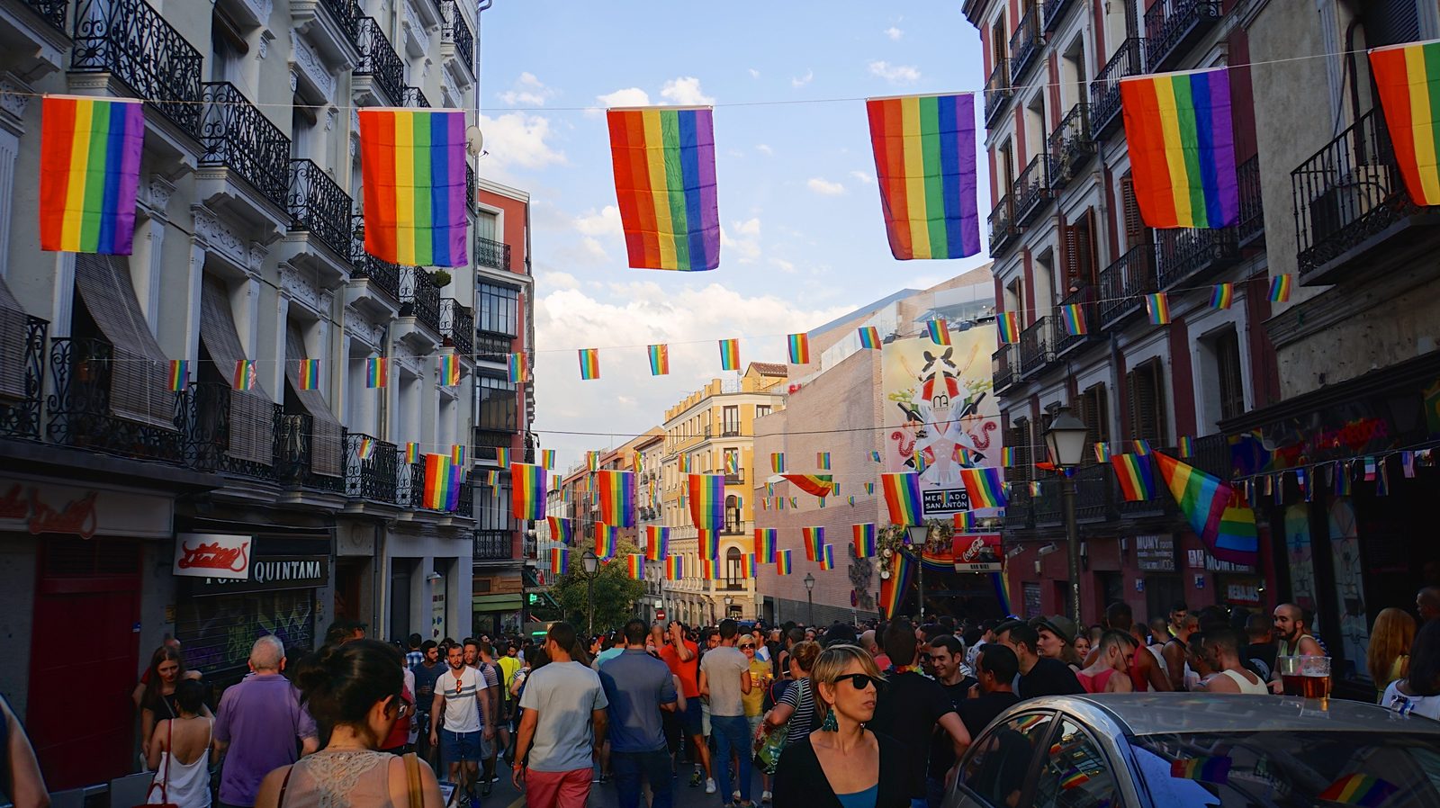 A crowded Chueca street strung with rainbow Pride flags between the balconied apartment buildings