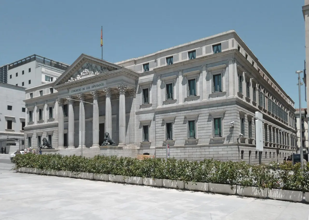 The Neoclassical facade of the Congreso de los Diputados in Madrid, with its columned portico, triangular pediment and two bronze lions flanking the entrance stairway under a clear blue sky