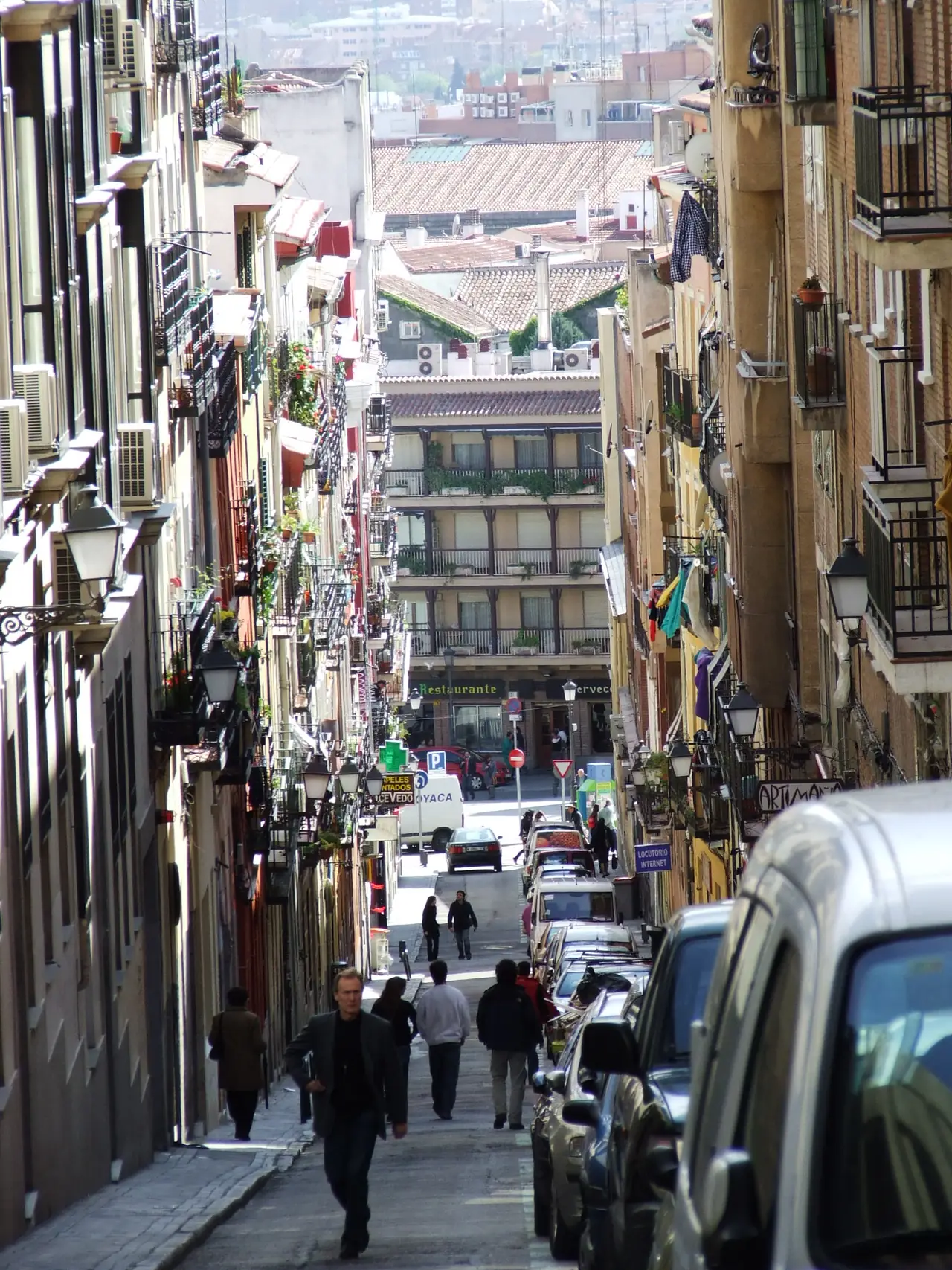 A narrow sloping street in Lavapiés lined with balconied apartment buildings, parked cars, and pedestrians walking downhill