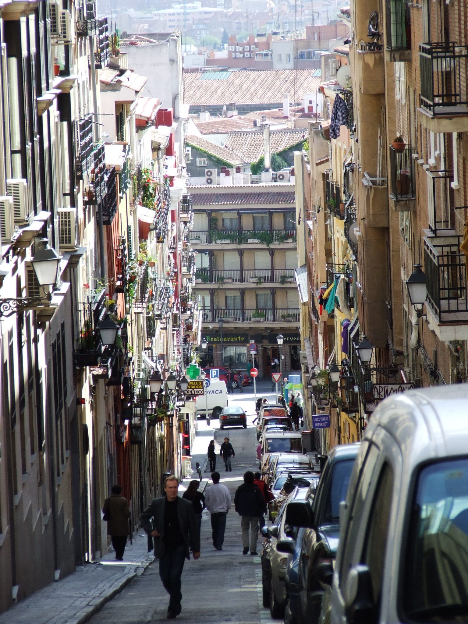 A narrow sloping street in Lavapiés lined with balconied apartment buildings, parked cars, and pedestrians walking downhill