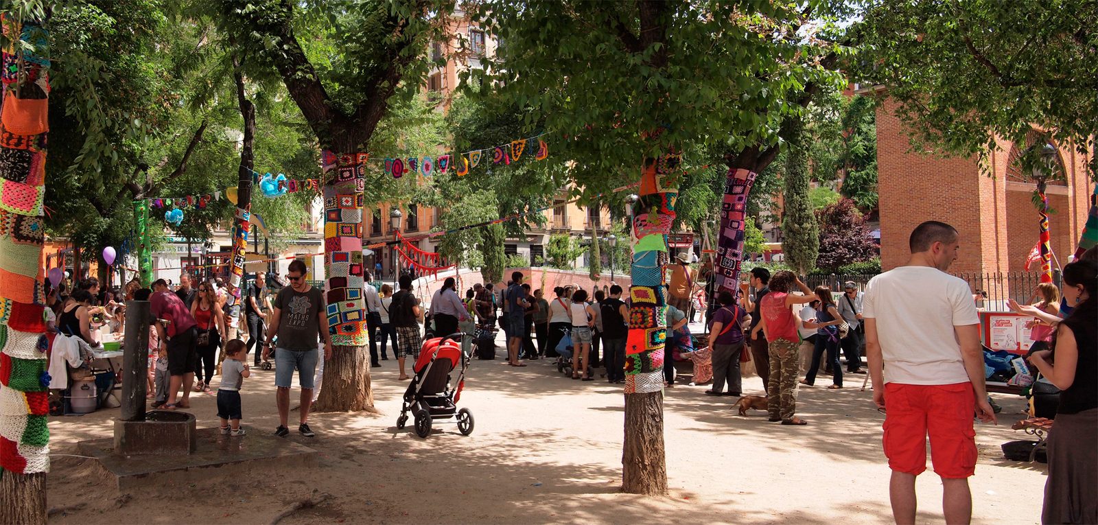 A tree-lined plaza in Malasaña with tree trunks wrapped in colorful yarn bombing and a crowd gathered for a street event