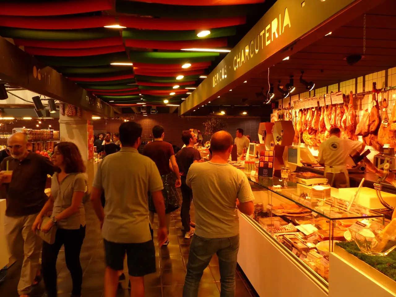 Inside Mercado de San Antón in Madrid, an Iberian charcuterie counter with hanging jamones and a carver at work as shoppers browse the gourmet market hall