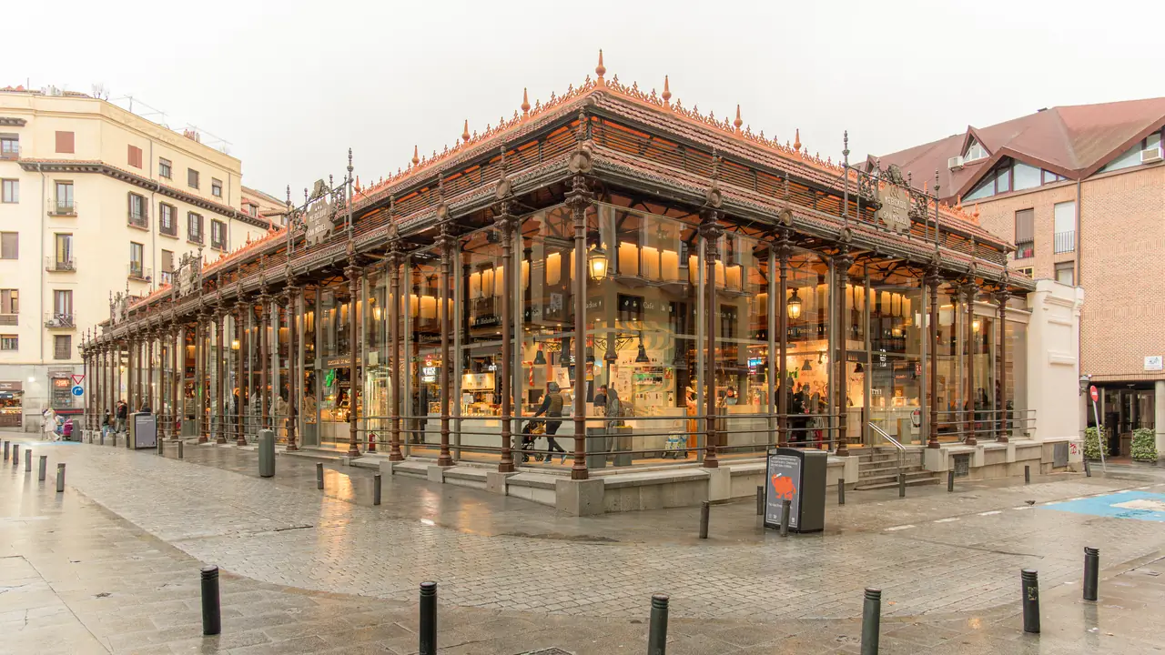 The glass-and-ironwork exterior of Mercado de San Miguel lit up at dusk, with its 'Mercado de San Miguel' signage on the roofline
