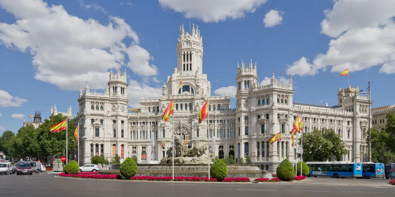 The white stone facade and central clock tower of the Palacio de Cibeles, with Spanish flags flying and the Cibeles fountain in front