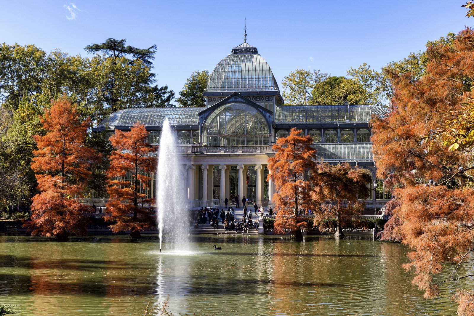The glass-domed Palacio de Cristal reflected in its pond behind a fountain jet, framed by orange autumn foliage in Retiro Park