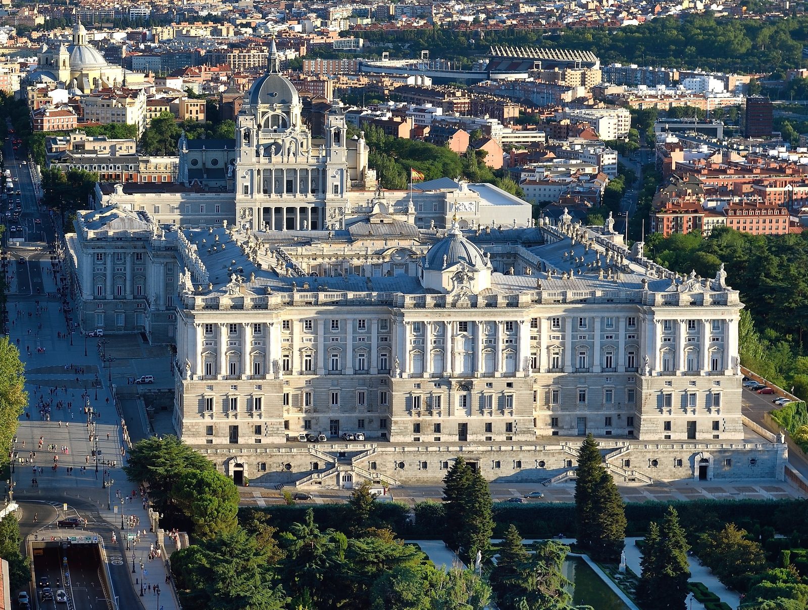 Aerial view of the Royal Palace of Madrid's stone facade and courtyards, with the Almudena Cathedral's dome behind it