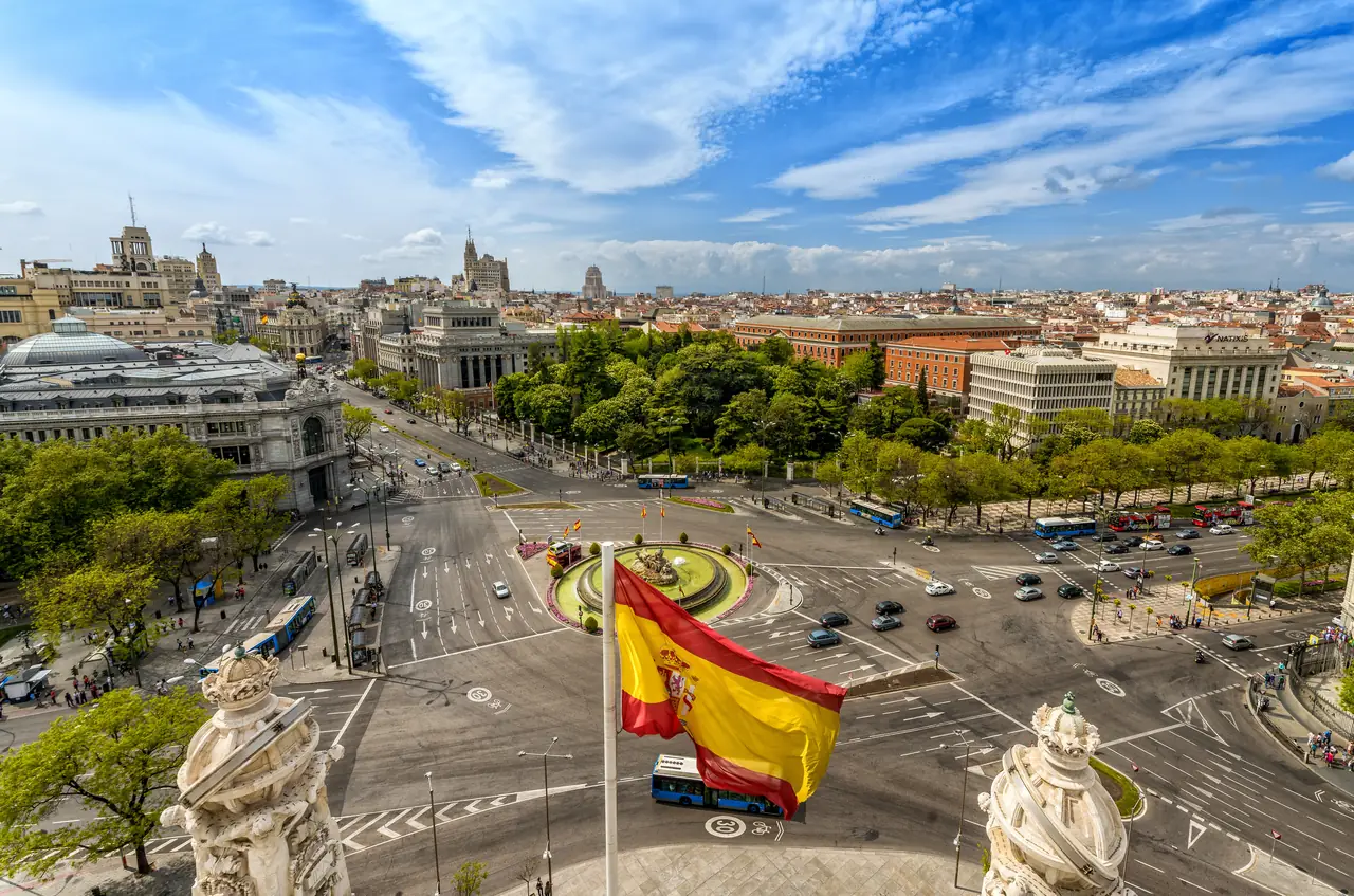 Aerial view of the Cibeles fountain roundabout at the center of Plaza de Cibeles, with a Spanish flag flying in the foreground and traffic circling below