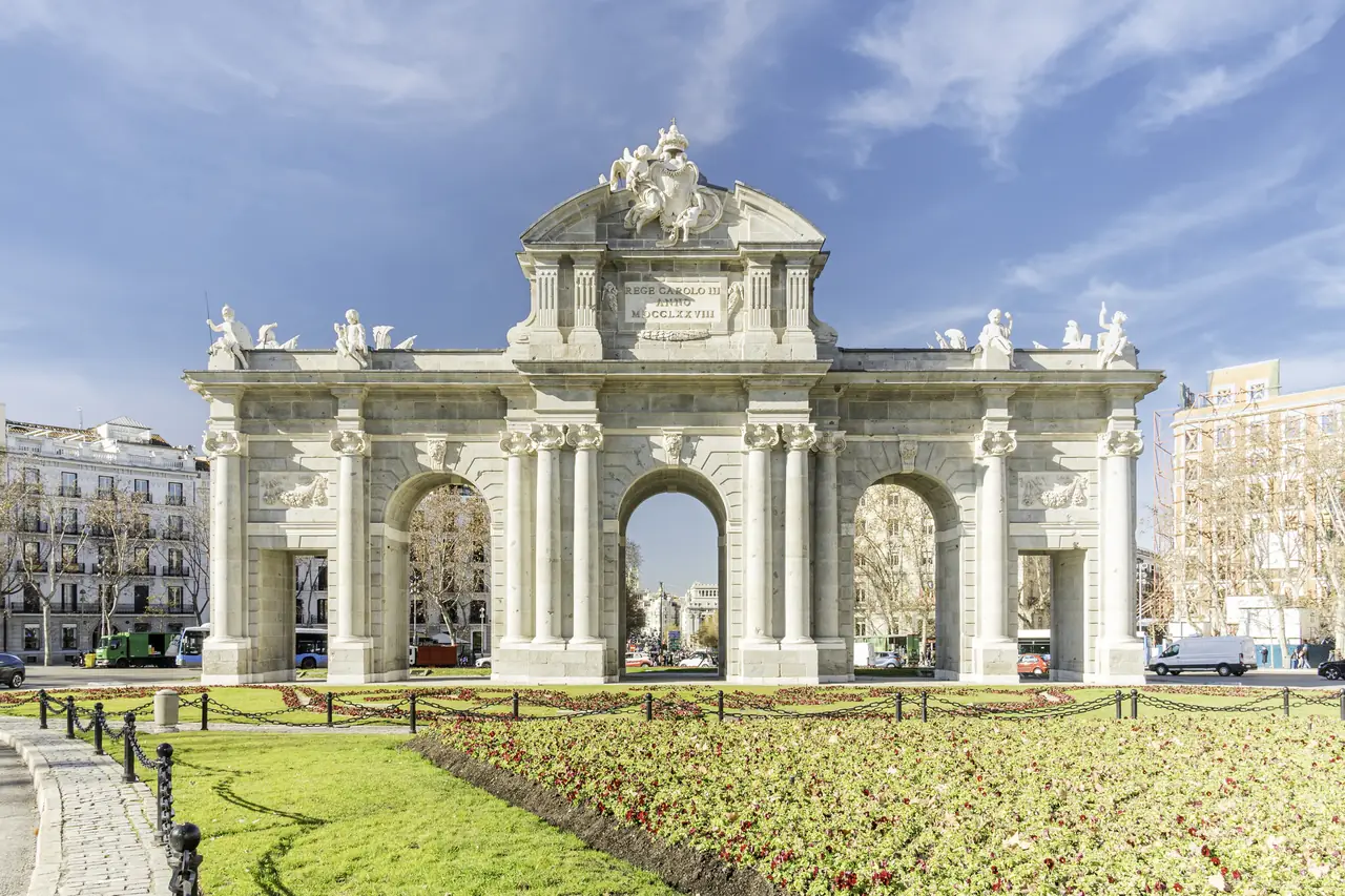 The stone triple-arched Puerta de Alcalá viewed head-on, with its carved crest and inscription against a blue sky