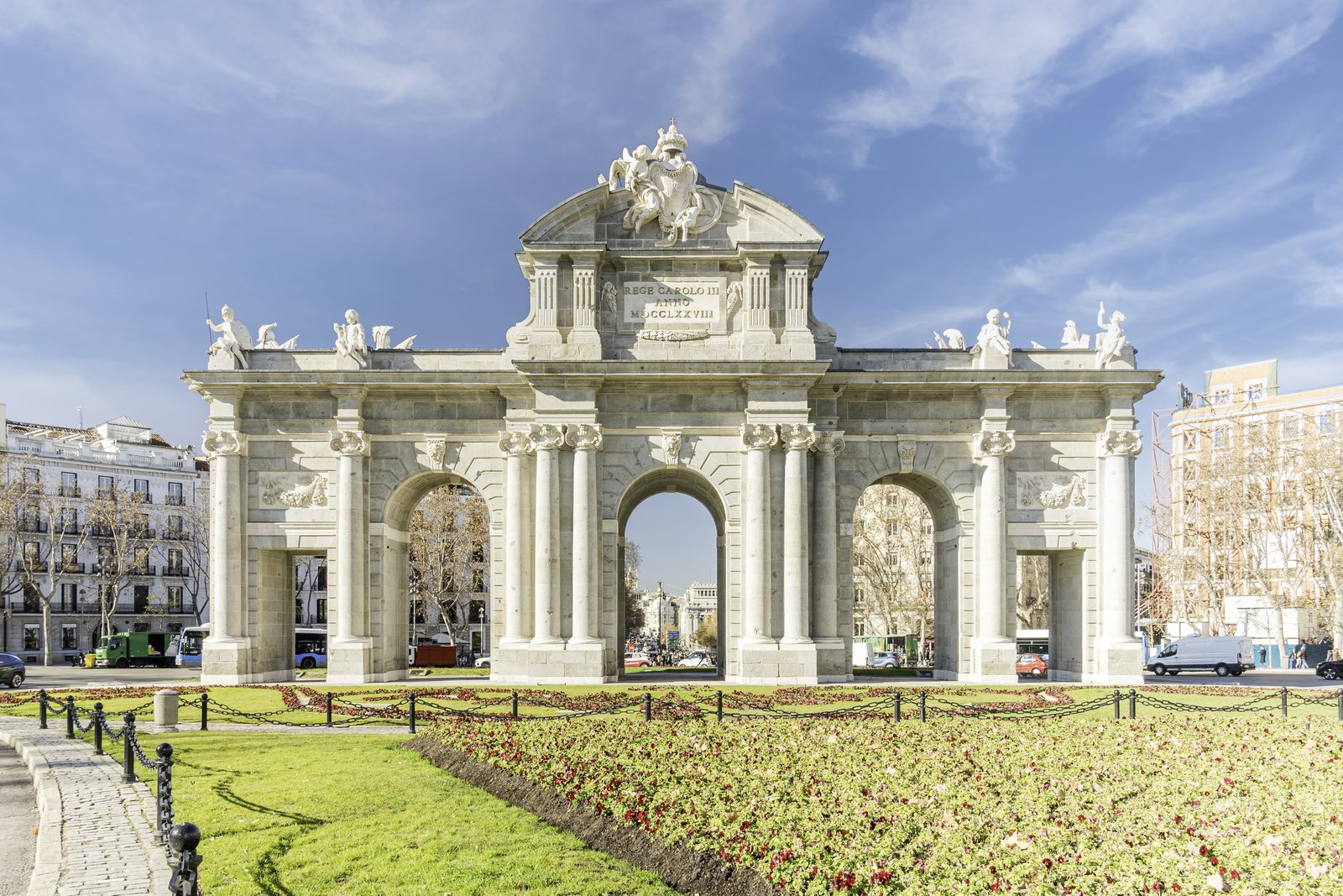 The stone triple-arched Puerta de Alcalá viewed head-on, with its carved crest and inscription against a blue sky