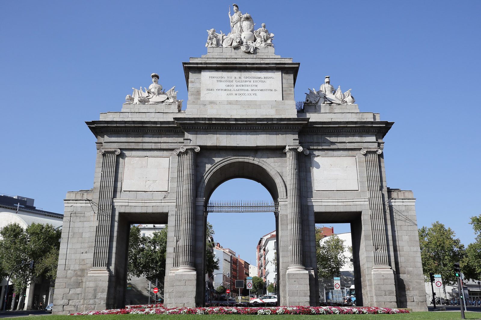 The Puerta de Toledo in Madrid, a Neoclassical stone gateway with a central arch, flanking columns and sculptural groups crowning the top, set among flowerbeds under a blue sky
