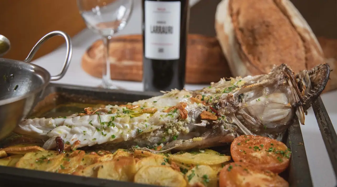 Whole roasted fish on a metal tray with sliced potatoes and grilled tomato, a bottle of Larrauri wine and rustic bread behind