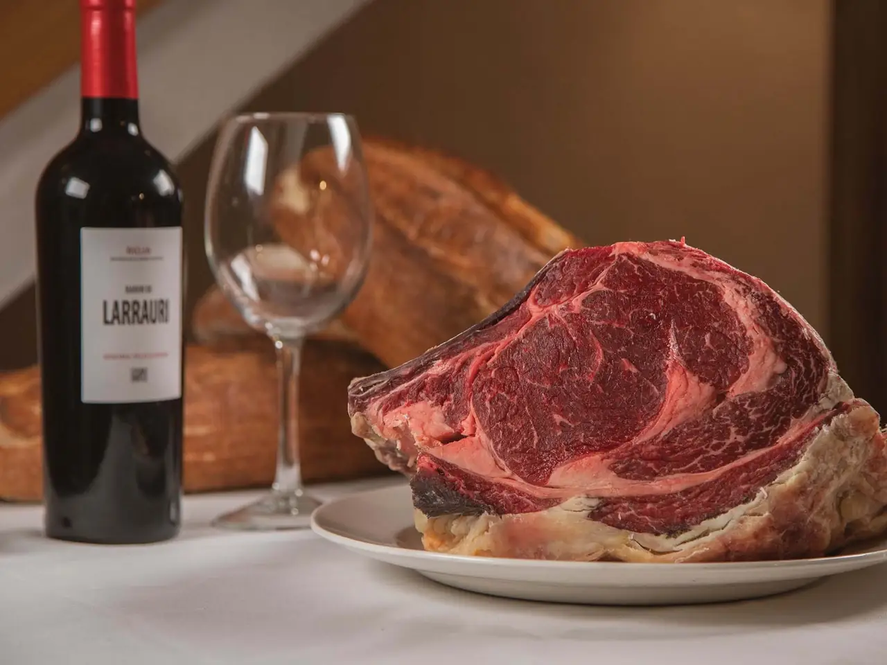 Large bone-in rib of marbled raw beef on a white plate beside a bottle of Baron de Larrauri Rioja and rustic bread