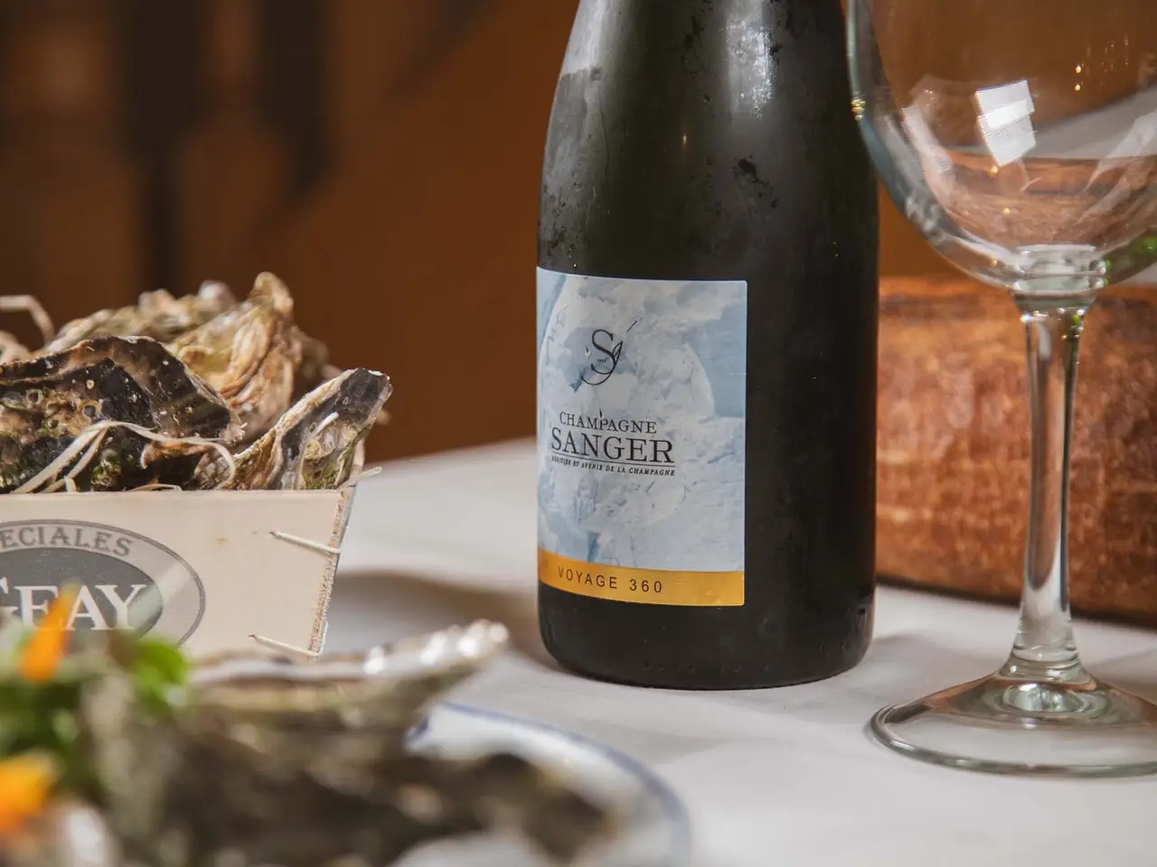 Table setting with a crate of fresh oysters, a chilled bottle of Champagne Sanger and a wine glass on white linen