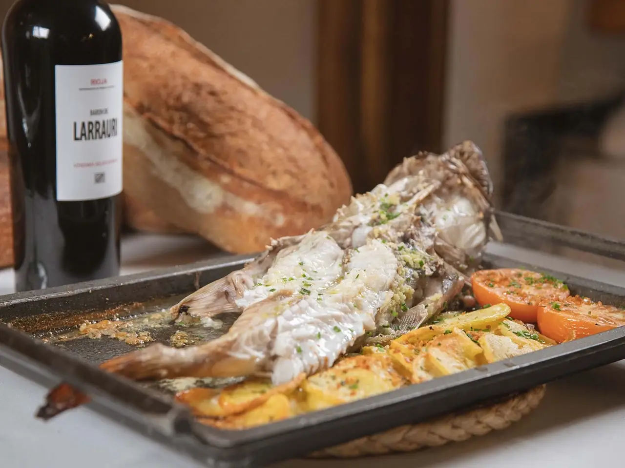Whole baked fish on a tray with roast potatoes and grilled tomato, a bottle of Baron de Larrauri wine and bread behind