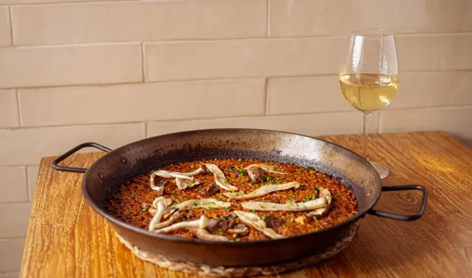 Wild mushroom paella in a wide black paella pan on a wooden table, with a glass of white wine beside it, at L'Arrosseria Xàtiva (Gràcia)