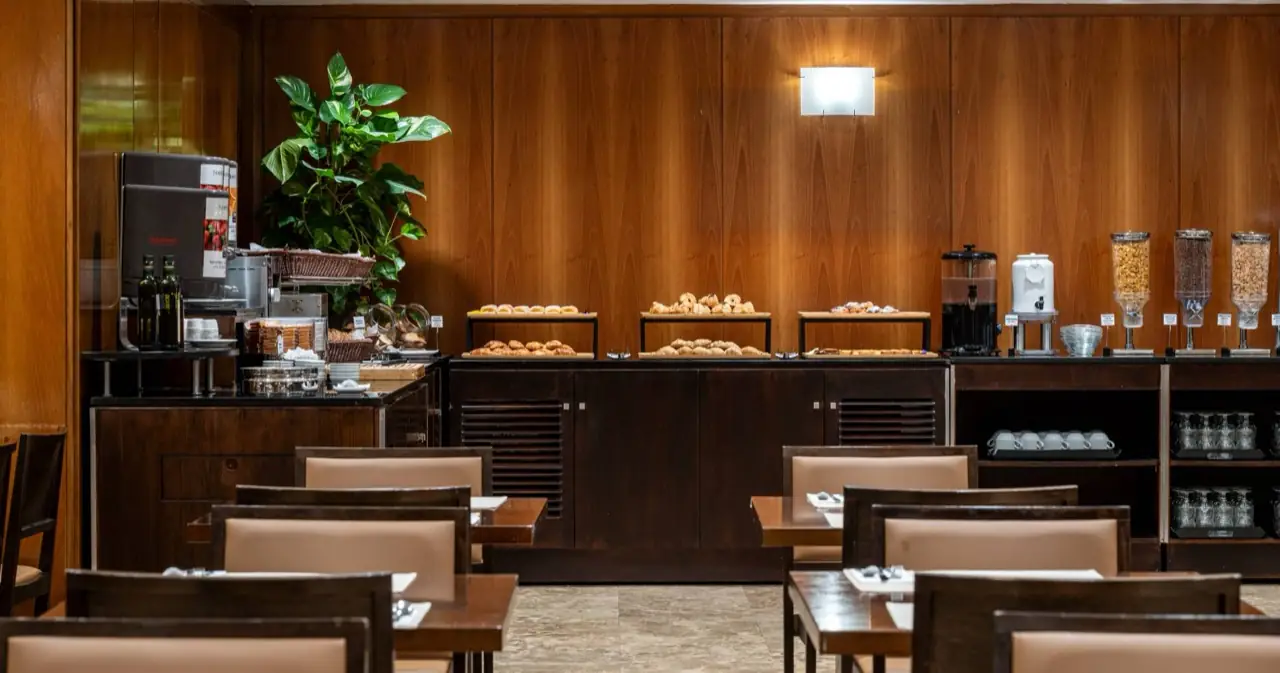 Breakfast buffet room with trays of pastries and croissants laid out on a wood-paneled counter beside set dining tables