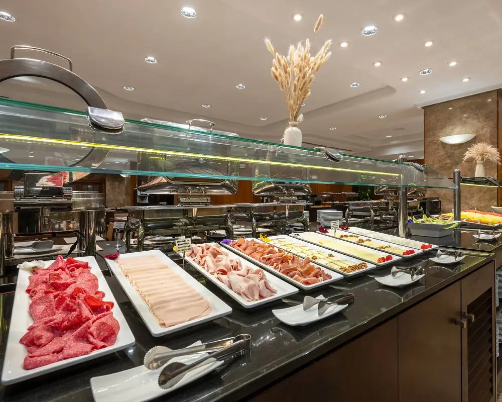 Buffet counter with trays of sliced cured meats, ham and cheese under a glass sneeze guard