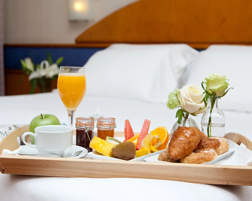 Wooden breakfast tray on a bed holding orange juice, coffee, croissants, jams and fresh fruit