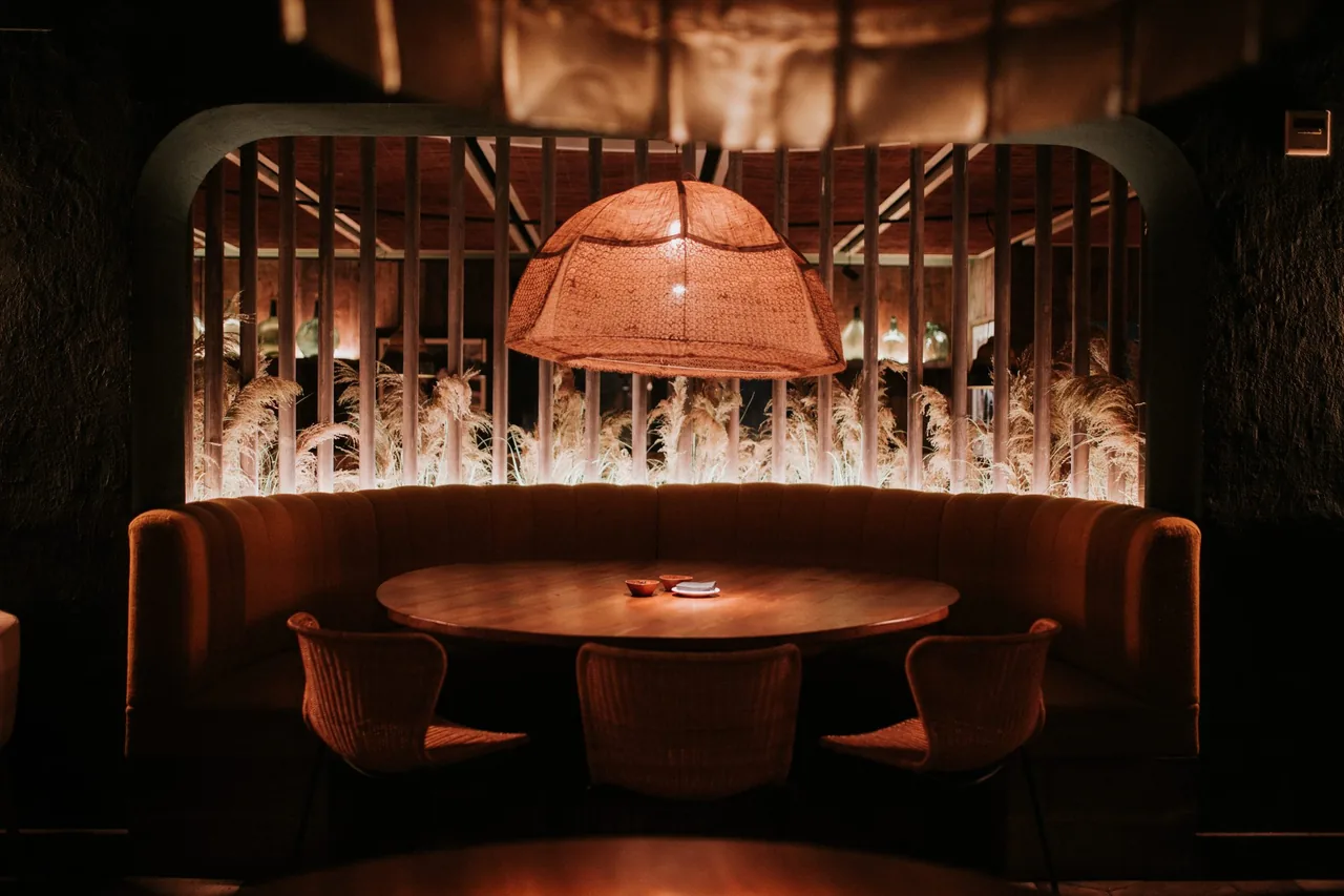 Warmly lit dining nook with a curved velvet banquette around a round wooden table, a large woven dome pendant overhead, and backlit pampas grass behind a slatted timber screen