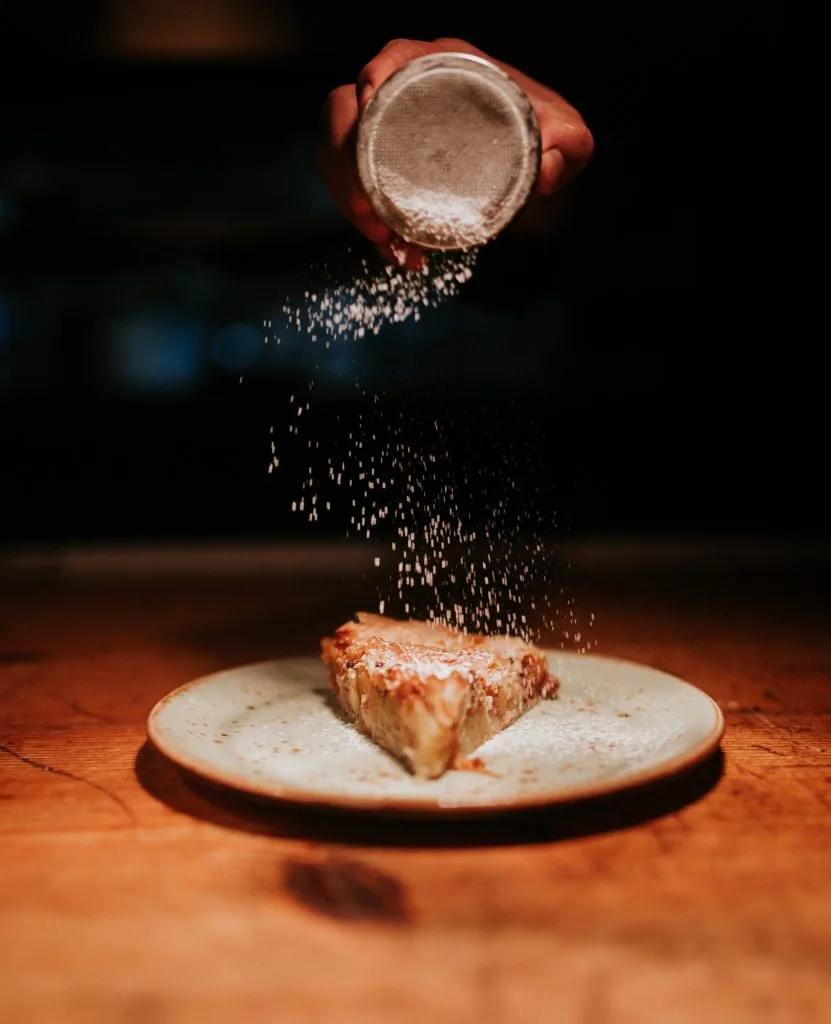 A hand shaking icing sugar from a sieve over a wedge of golden tart on a speckled ceramic plate on a dark wooden table