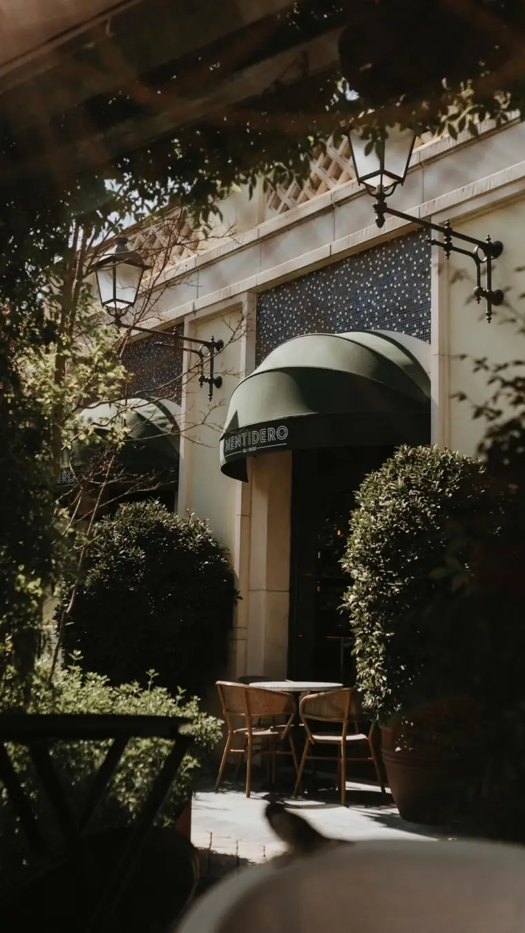 Exterior of Mentidero restaurant with a green striped awning, wall lanterns, and an outdoor table with rattan chairs framed by greenery
