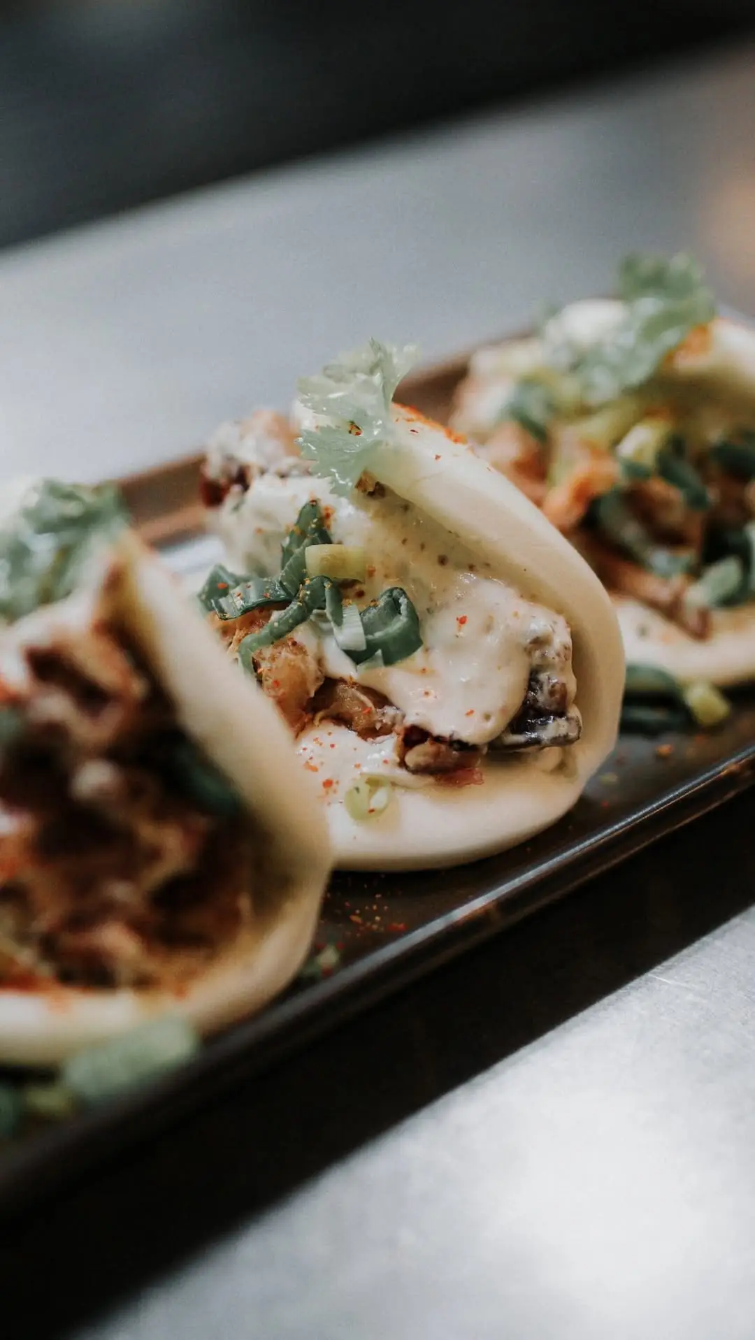 Close-up of steamed bao buns filled with fried shrimp, pickles, and scallion on a dark tray