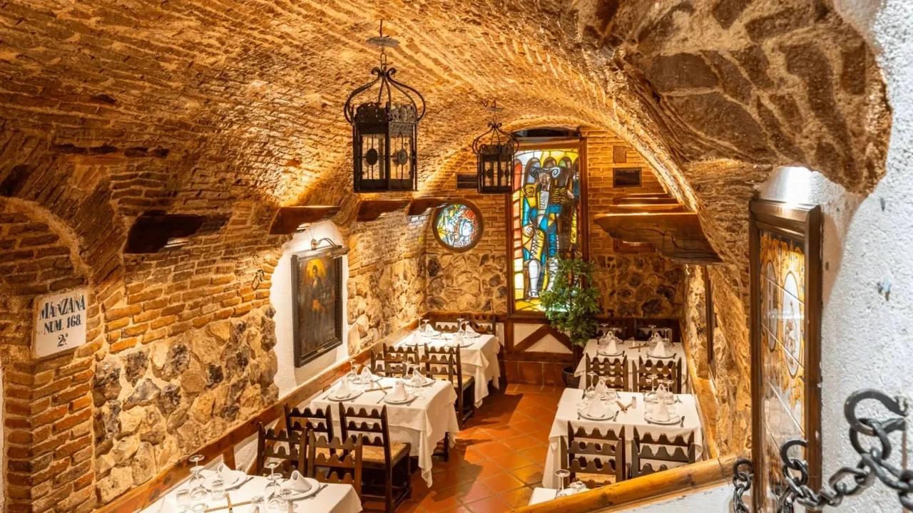 A vaulted stone and brick cellar dining room with tables set with white cloths and stained glass on the wall.