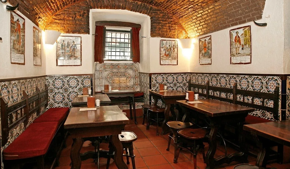 A dining room with a tiled lower wall, a red bench, wooden tables and a brick vaulted ceiling.