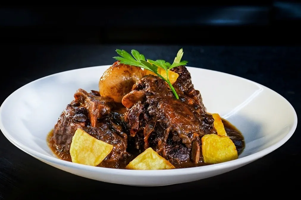 A white bowl of braised meat with potatoes and a sprig of herb on a dark table