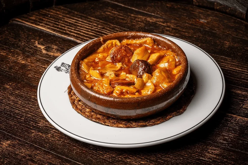 A clay cazuela of a tomato-red stew on a white plate on a wooden table