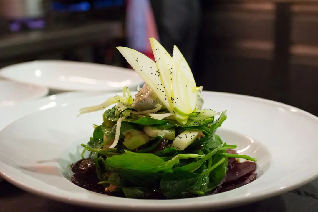 Layered green salad with arugula, thin apple slices, and poppy seeds on a white plate, Las Fernández.