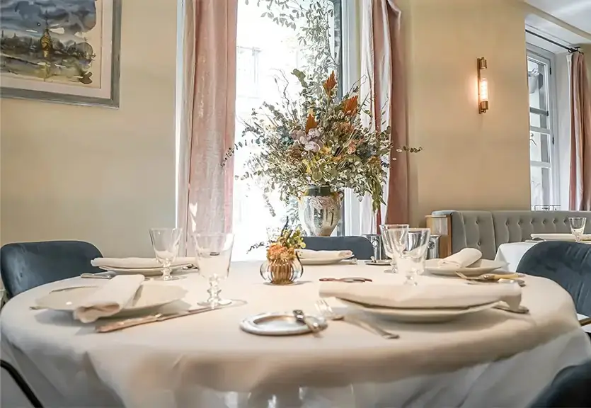 Round dining table set with white linen, glassware and a tall dried-flower arrangement by a curtained window, blue velvet chairs around it