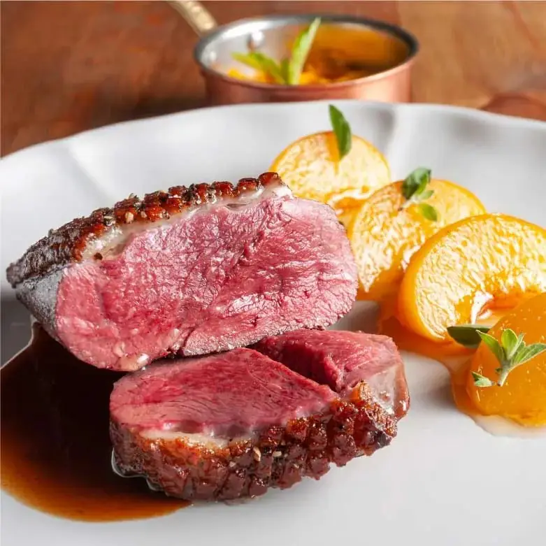 Sliced rare seared duck breast in dark jus with roasted orange segments and a copper saucepan behind, on a white plate