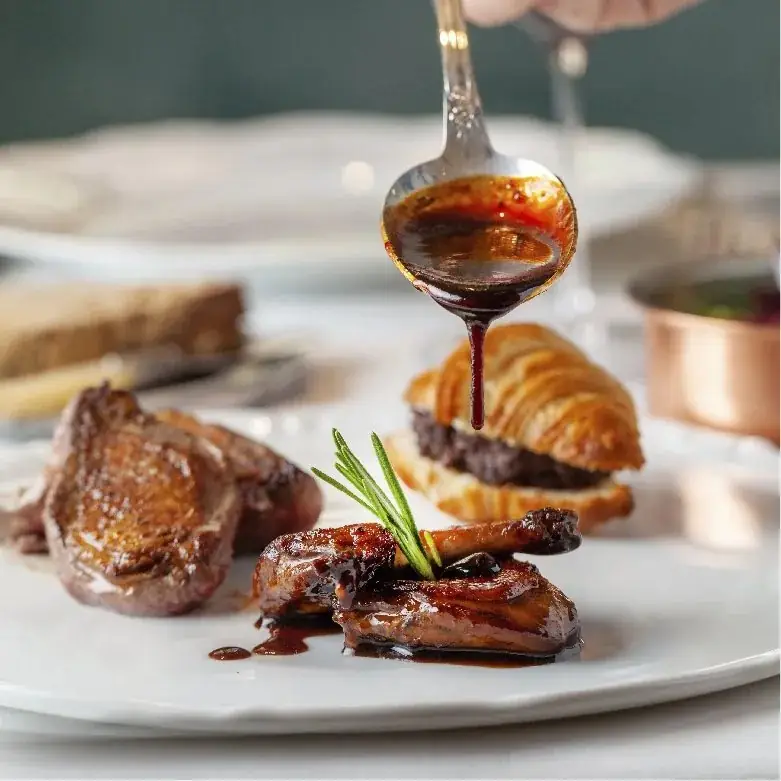 Spoon pouring dark jus over a plated dish of seared meat, a croissant with confit filling and rosemary garnish