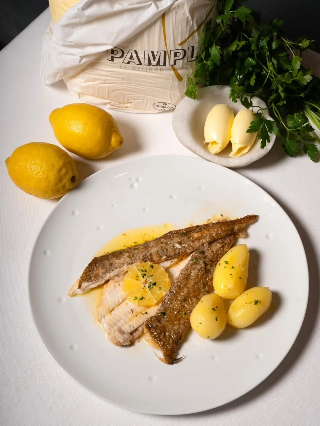 A white plate with a fried fish fillet and boiled potatoes with lemon and butter beside it