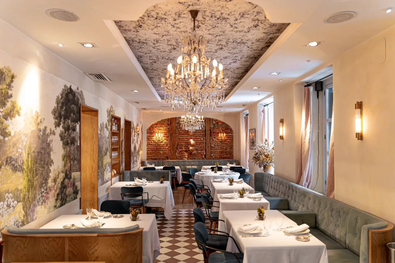 A long dining room with a chandelier, painted ceiling, white-clothed tables and a red brick wall at the far end
