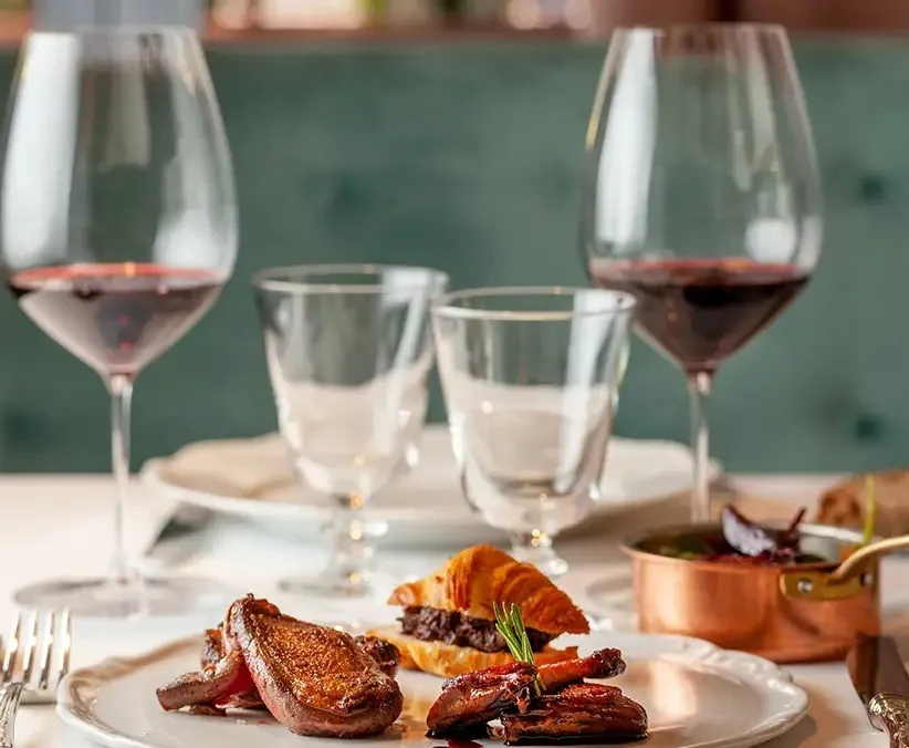Two glasses of red wine beside a plate of meat with a small copper pot