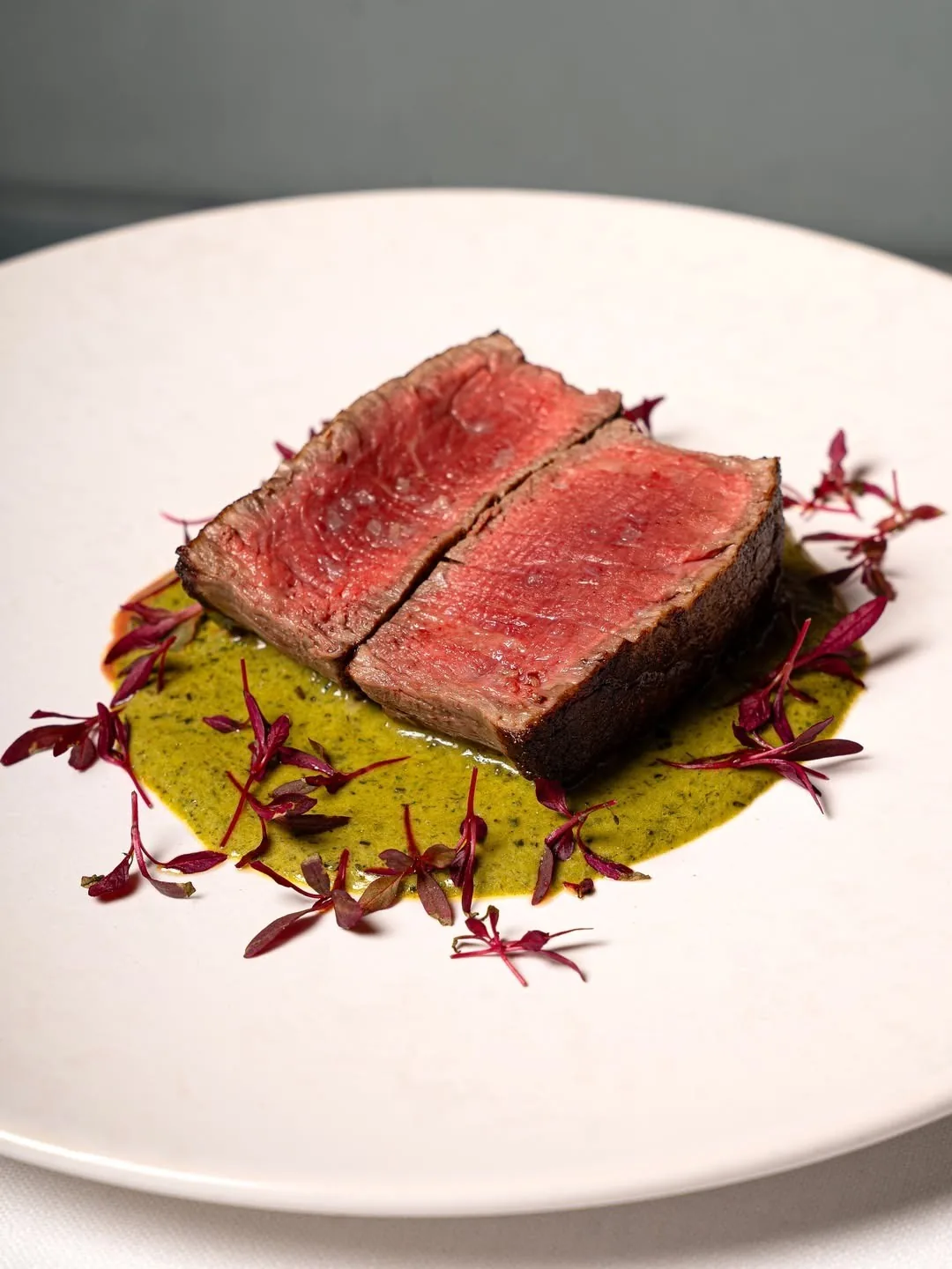 Two slices of rare seared beef on a white plate over a green sauce with red leaves