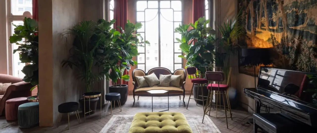 Ornate lounge interior with large potted plants, antique upholstered seating, a piano and a tapestry on the wall
