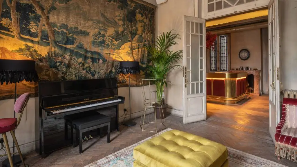 Ornate lounge room with an upright piano, tapestry-covered wall, and a golden velvet ottoman, opening onto a gilded bar area