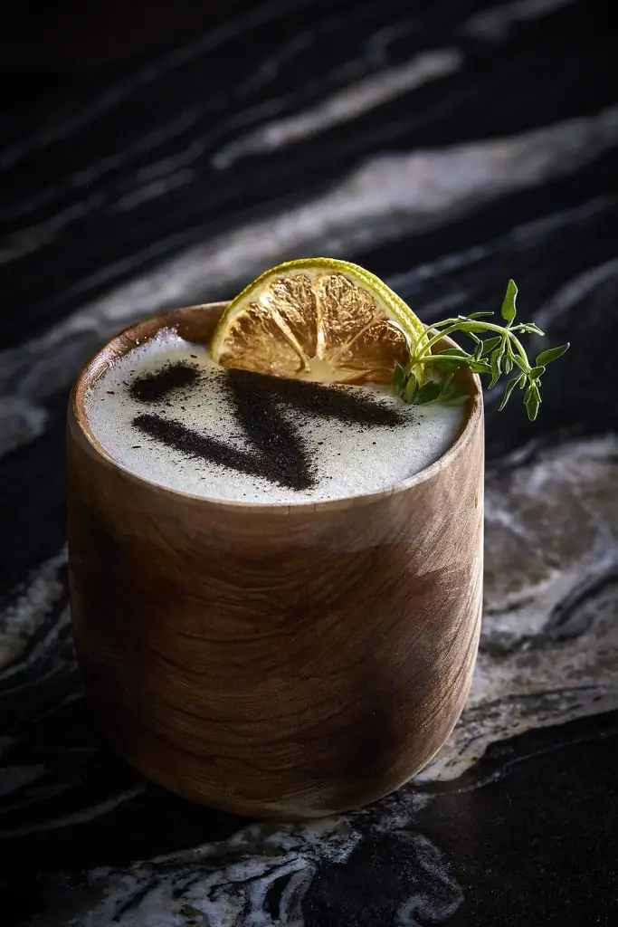 Frothy cocktail in a wooden cup garnished with a dried citrus wheel and thyme, topped with a dark powder motif, on marble