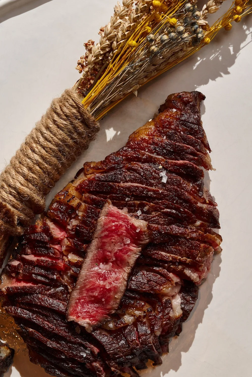 A grilled steak with a slice cut from it on a board beside a bundle of dried herbs