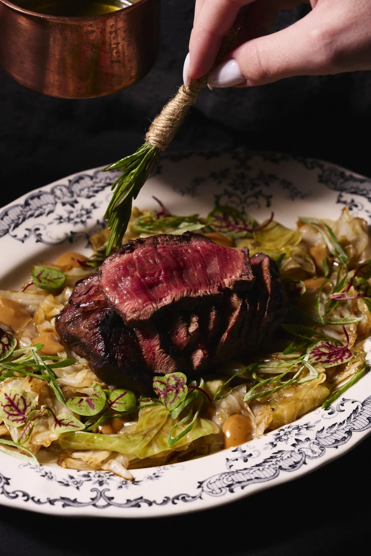 A seared slice of beef on a patterned plate with vegetables, a hand placing a herb sprig