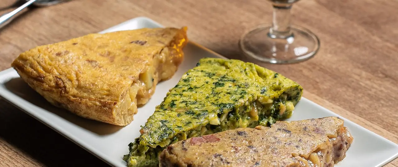 Close-up of three sliced Spanish tortillas — potato, spinach, and a savory pâté-like variety — plated on a white rectangular dish, Les Truites
