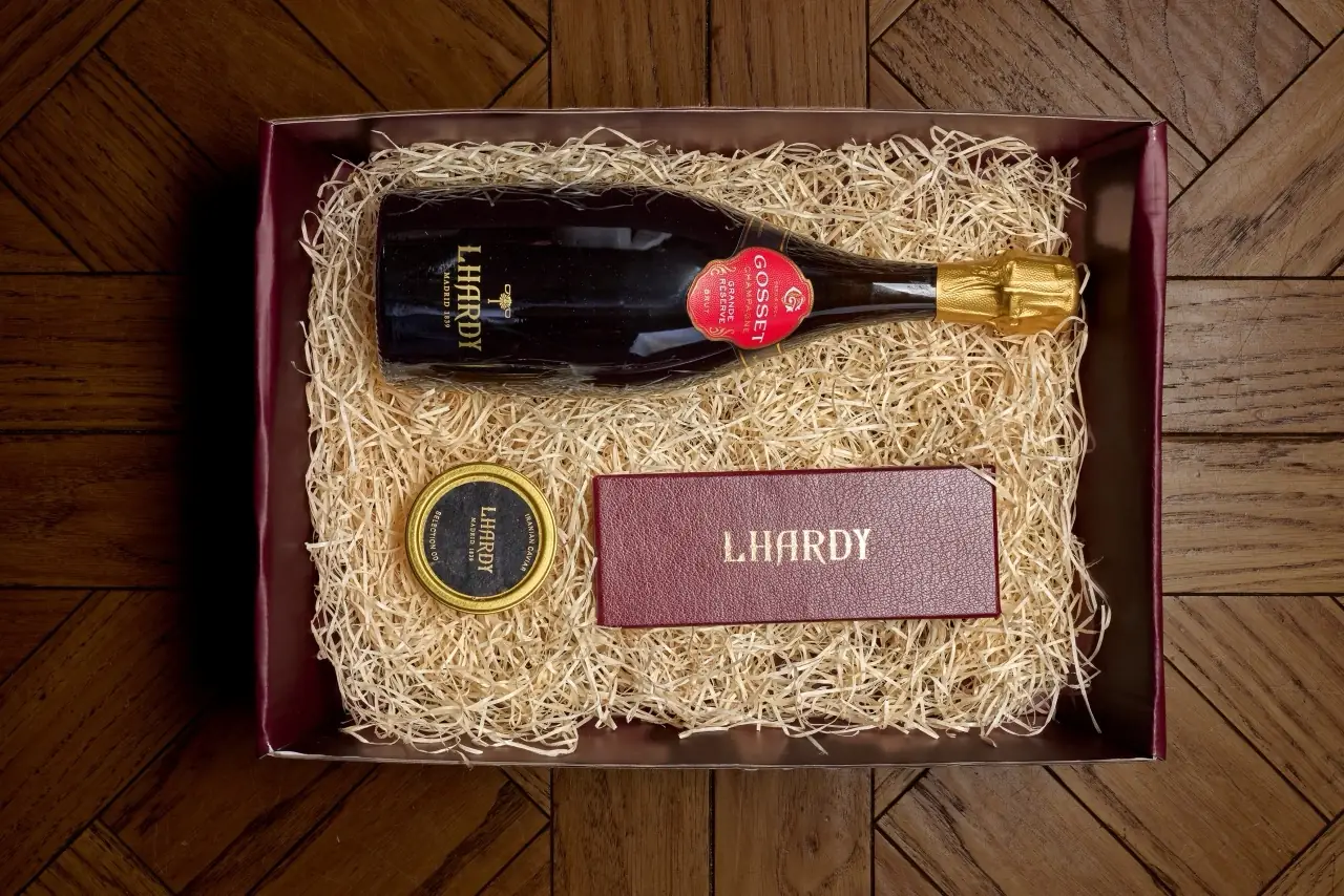 Overhead gift box lined with wood-wool straw holding an Lhardy-labelled Gosset champagne bottle, a gold-lidded tin of Iranian caviar and a burgundy leather 'Lhardy' menu card, on a parquet floor