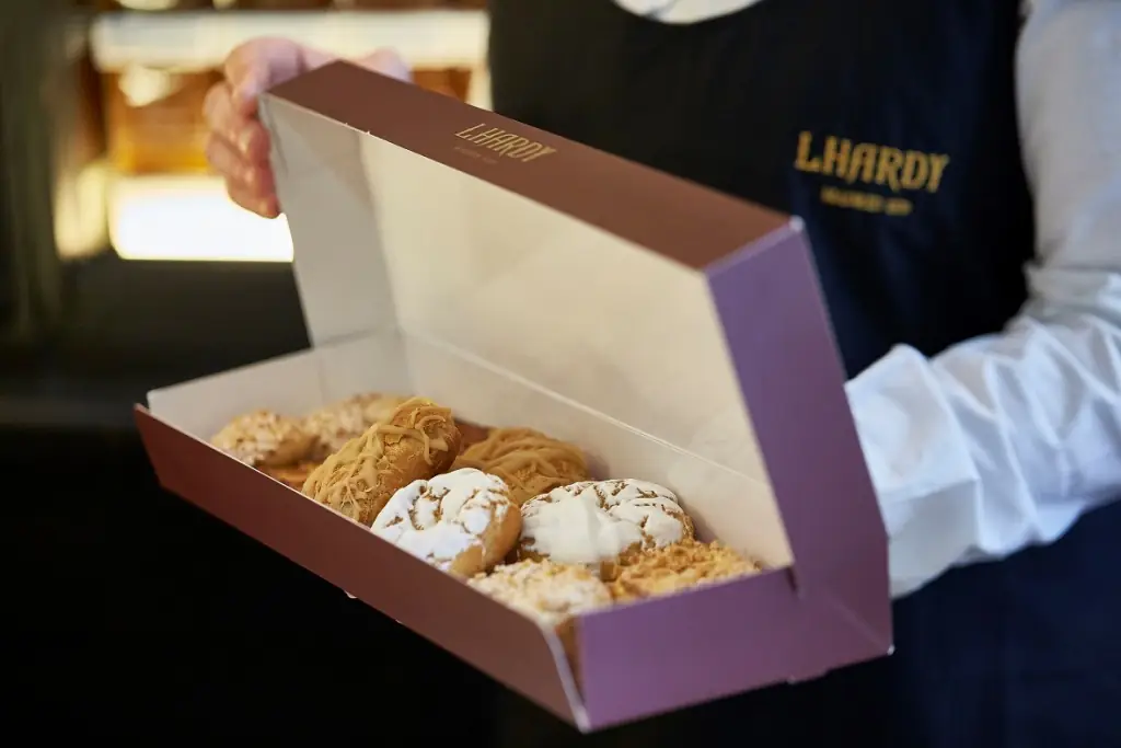 Open burgundy pastry box held by a staff member in an 'Lhardy'-embroidered navy apron, filled with assorted glazed and iced traditional pastries