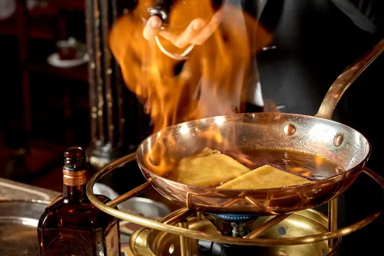 Tableside flambe in a copper pan set over a burner ring, flames rising over folded crepes in dark sauce, a waiter pouring liquor and a bottle beside the stand