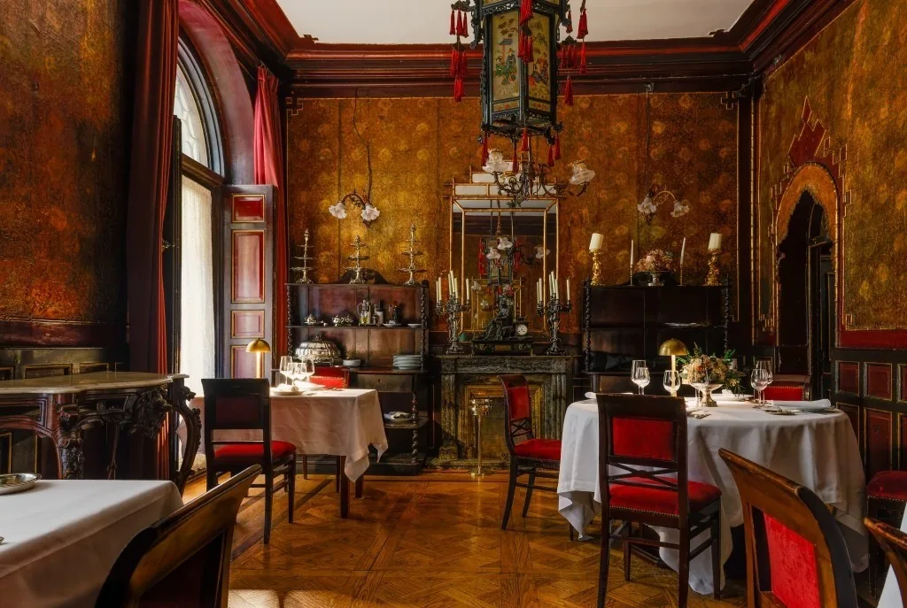A dining room with red upholstered chairs, white tablecloths, gilded wall panels and a hanging lantern
