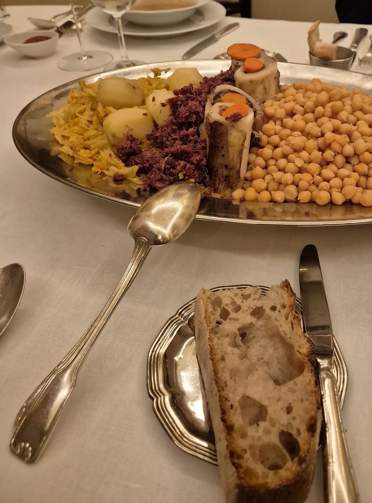 A silver platter of cocido chickpeas, cabbage, potatoes and meats, with bread and a serving spoon