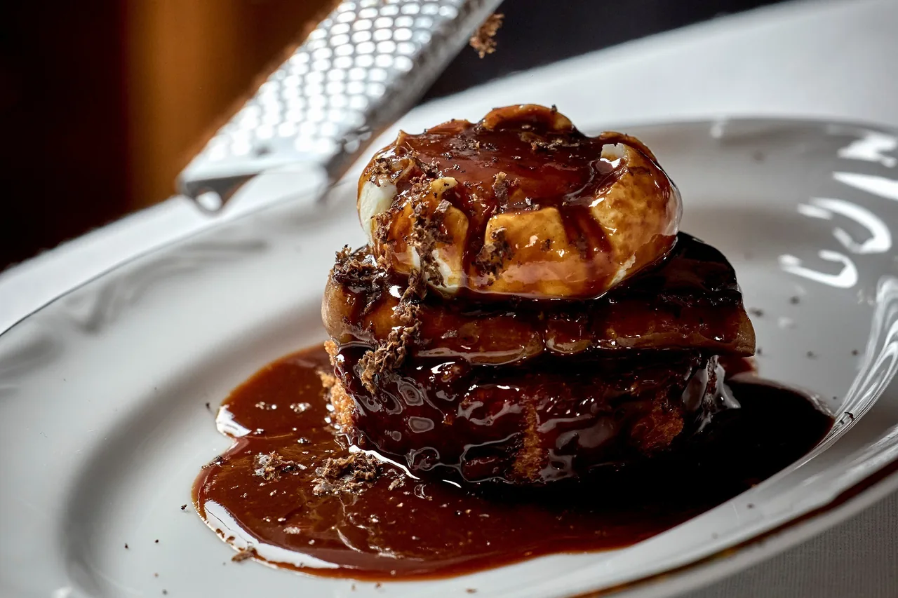 A stacked dish glazed with dark sauce on a white plate with an embossed rim